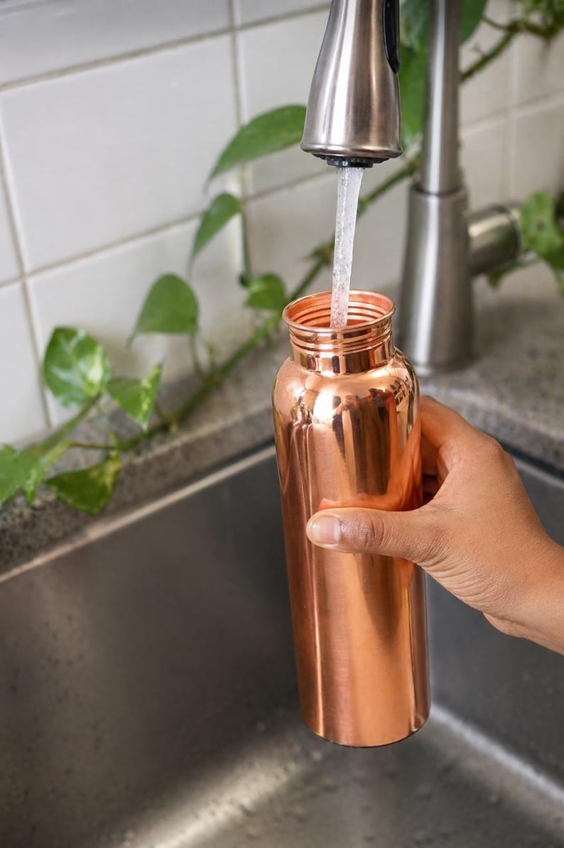 May include: A copper water bottle being filled with water from a stainless steel faucet. The bottle is cylindrical with a narrow neck. The background includes a stainless steel sink, white tile, and green leafy plants.