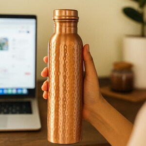 Puede incluir: Una botella de agua de color cobre con un dise&ntilde;o texturizado y martillado. La botella est&aacute; sujeta por una mano. Tiene una tapa de rosca y forma cil&iacute;ndrica. El fondo incluye un port&aacute;til y una planta.