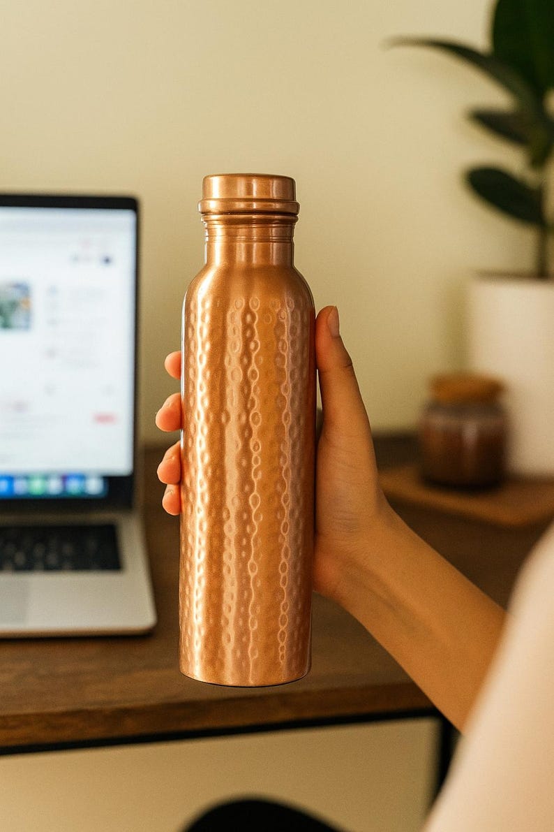 Puede incluir: Una botella de agua de color cobre con un dise&ntilde;o texturizado y martillado. La botella est&aacute; sujeta por una mano. Tiene una tapa de rosca y forma cil&iacute;ndrica. El fondo incluye un port&aacute;til y una planta.