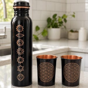 K&ouml;nnte beinhalten: Eine schwarze Metallwasserflasche mit kupferfarbenem Schraubverschluss und sieben vertikalen Chakrasymbolen in Kupfer, begleitet von zwei passenden schwarzen Metallbechern mit kupferfarbenem Innenraum und geometrischem Lebensblumenmuster.