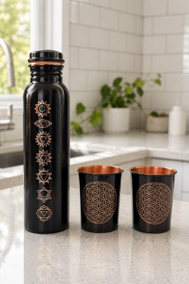 K&ouml;nnte beinhalten: Eine schwarze Metallwasserflasche mit kupferfarbenem Schraubverschluss und sieben vertikalen Chakrasymbolen in Kupfer, begleitet von zwei passenden schwarzen Metallbechern mit kupferfarbenem Innenraum und geometrischem Lebensblumenmuster.