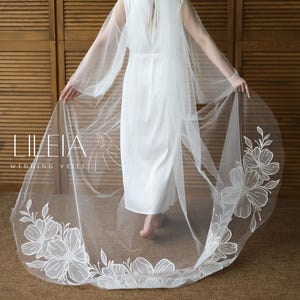 May include: A bride in a white wedding dress and veil. The veil is long and sheer, with a floral design along the edges. The text "LILEIA WEDDING VEILS" is visible in the lower left corner. The background is a wooden wall.