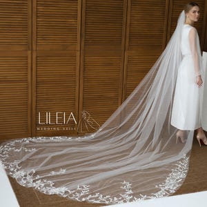 May include: A long, flowing white wedding veil with delicate floral lace trim. The veil is paired with a white dress. The image also features the text "Lileia Wedding Veils".