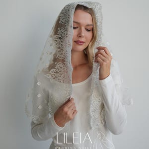 Puede incluir: Un velo de novia blanco transparente con intrincados detalles de encaje floral. El velo está drapeado sobre la cabeza y los hombros, mostrando el delicado patrón. Incluye el texto "LILEIA WEDDING VEILS".