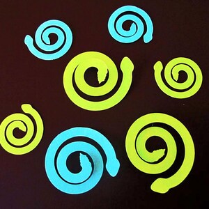 May include: A collection of paper cut-out spirals in shades of light blue and neon yellow-green, arranged on a dark brown background. The spirals vary in size and are scattered across the surface.