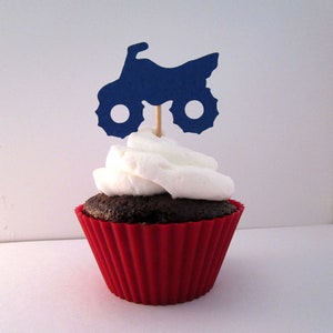 May include: A chocolate cupcake with white frosting topped with a blue paper silhouette of a dirt bike.
