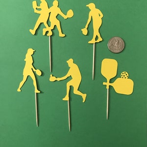 Pickleball Players in silhouette are cut from yellow cardstock against a background of green. Cutouts in various action poses and are attached to wooden picks. A US quarter is shown for size comparison.
