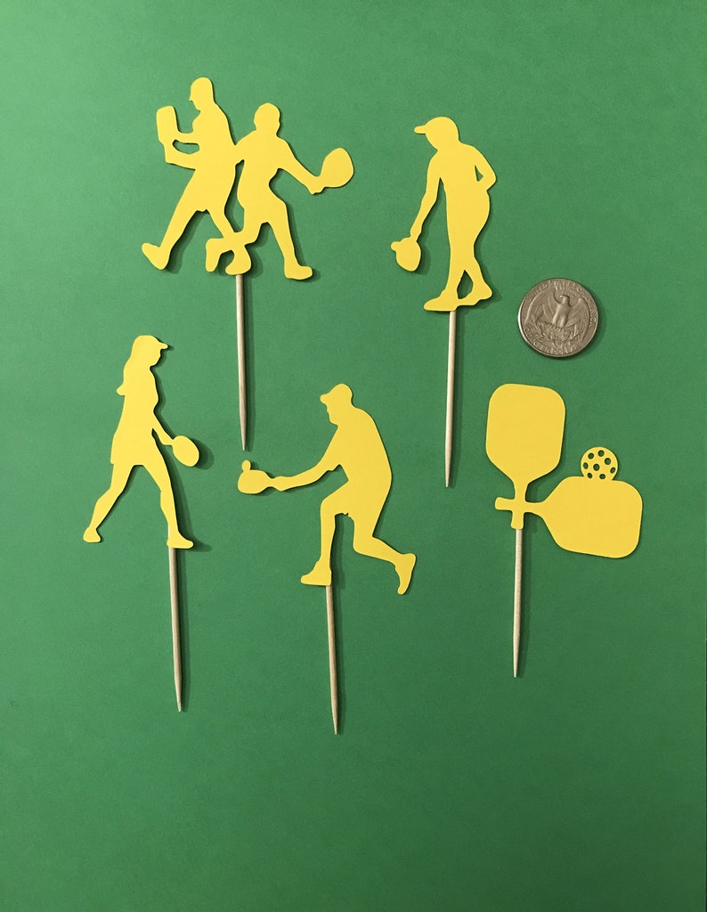 Pickleball Players in silhouette are cut from yellow cardstock against a background of green. Cutouts in various action poses and are attached to wooden picks. A US quarter is shown for size comparison.