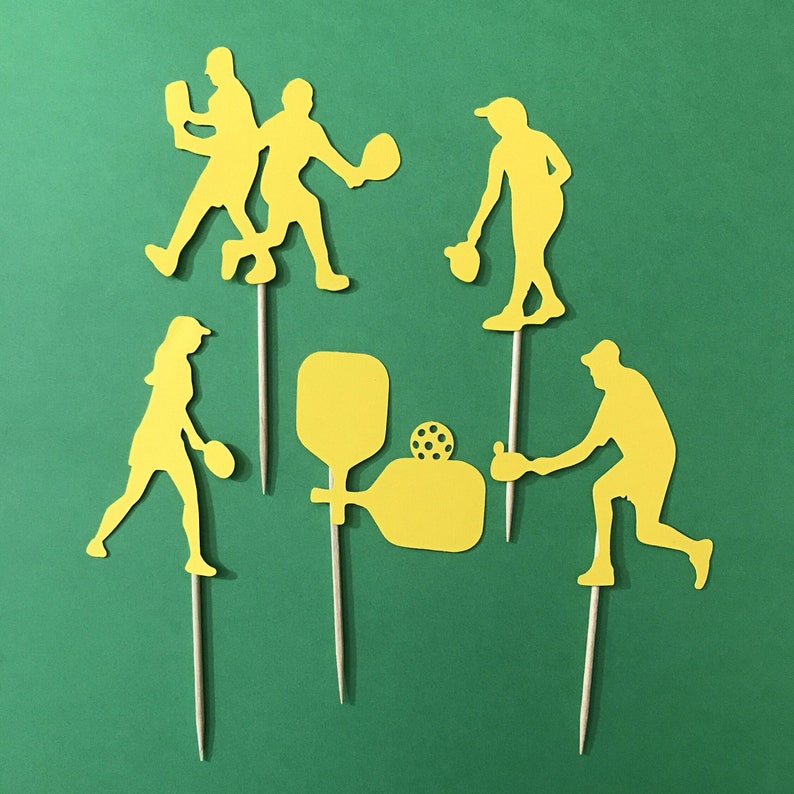 Pickleball Players in silhouette cut from yellow cardstock against a background of green. Silhouettes are attached to wooden picks to be used as Cupcake Toppers or food picks. Players are shown holding pickleball paddles, pickleball paddles with ball