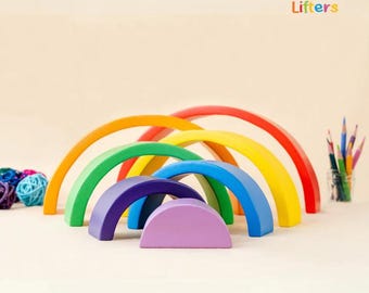 Könnte beinhalten: Ein Holz-Regenbogen-Stapelspielzeug in Rot, Orange, Gelb, Grün, Blau und Lila. Das Spielzeug besteht aus Bögen in verschiedenen Größen, ideal für kreatives Spielen und Lernen. Das Little Lifters Logo ist im Bild zu sehen.