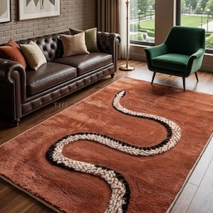 Puede incluir: Una alfombra rectangular de color naranja terracota profundo que presenta un prominente motivo de serpiente sinuosa. El diseño de la serpiente está realizado en un pelo texturizado y elevado con un patrón de segmentos negros y crema. El texto The Kaleen Rugs es visible.