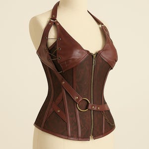 K&ouml;nnte beinhalten: Ein braunes Korsett mit Neckholder-Design. Es hat einen Frontrei&szlig;verschluss, dekorative Riemen und einen G&uuml;rtel mit gro&szlig;em Ring. Das Oberteil hat ein Blumenmuster und besteht aus einer Materialkombination.