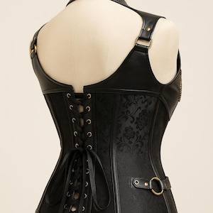 K&ouml;nnte beinhalten: Schwarzes Korsett mit floralem Muster und Lederakzenten. Das Korsett hat einen Neckholder, Schultertr&auml;ger und eine Schn&uuml;rung am R&uuml;cken. Goldfarbene Metallringe und eine Schnalle erg&auml;nzen das Design.