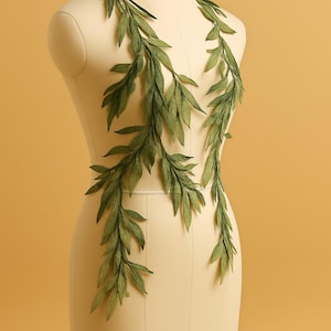 May include: Green leaf garland draped on a mannequin. The leaves are made of a textured, paper-like material, in various shades of green. The mannequin is a neutral beige colour, set against a warm yellow background. The garland is likely for decoration.