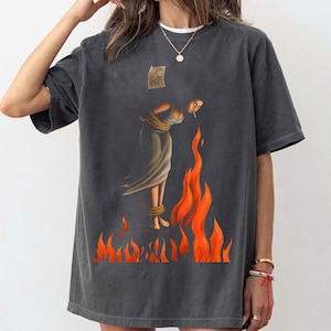 May include: Dark gray t-shirt featuring a surreal illustration of a person in a flowing dress, bound and engulfed in flames. The artwork includes a small, torn paper and is rendered in shades of gray, orange, and beige.