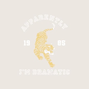May include: Cream-colored graphic with a yellow tiger design and black stripes. The text above the tiger reads "APPARENTLY". Below the tiger, the text reads "I'M DRAMATIC", with the numbers "19" and "85" on either side of the tiger.