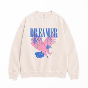 May include: A cream-coloured sweatshirt featuring the word "DREAMER" in blue block letters above a pink, blue, and green eagle graphic. The eagle is shown with outstretched wings, creating a striking visual on the front of the garment.