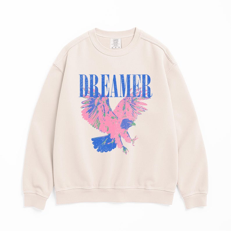 May include: A cream-coloured sweatshirt featuring the word "DREAMER" in blue block letters above a pink, blue, and green eagle graphic. The eagle is shown with outstretched wings, creating a striking visual on the front of the garment.