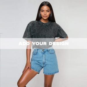 以下が含まれることがあります： 濃いグレーのアシッドウォッシュTシャツとライトブルーのデニムショーツを着た人。 Tシャツはクルーネックと半袖です。 ショーツはクラシックな5ポケットデザインです。 画像のテキストは「ADD YOUR DESIGN」です。
