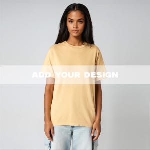 以下が含まれることがあります： 女性が、白文字で「ADD YOUR DESIGN」と書かれたマスタード色のTシャツを着ています。Tシャツは半袖で丸首です。彼女は水色のジーンズを履いています。背景は無地のライトグレーです。