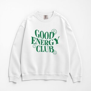 May include: White sweatshirt with the text "GOOD ENERGY CLUB" in green, along with smiley face graphics. The sweatshirt has a crew neck and long sleeves. The fabric appears to be a soft, comfortable material.