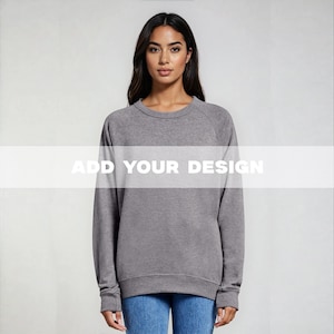 May include: A heather gray crew neck sweatshirt with long sleeves. The text "ADD YOUR DESIGN" is displayed across the front. The model is wearing blue jeans.
