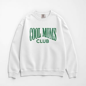 May include: White crew neck sweatshirt with the text "COOL MOMS CLUB" in distressed green lettering. The sweatshirt has a classic, casual style, perfect for everyday wear. The design is centered on the front.