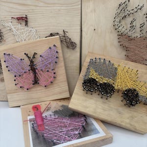 May include: A collection of string art projects on light wood boards, including a butterfly with pink and purple thread, a yellow and grey truck, a deer head, and a heart design with a spool of pink thread and a small bag of metal nails.