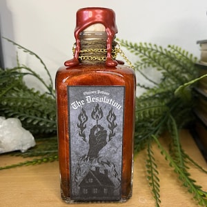 May include: A square glass bottle filled with a shimmering orange liquid. The bottle is sealed with a wooden cork and red wax. A label on the front reads "The Desolation" with a graphic of a hand holding a match over a burning house. The bottle is surrounded by greenery.