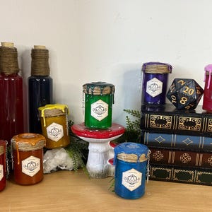 Könnte beinhalten: Eine Sammlung von Fantasy-Themen-Trankgläsern und -flaschen in verschiedenen Farben, darunter Rot, Orange, Gelb, Grün, Blau, Lila und Rosa. Jedes Glas hat eine einzigartige Farbe Wachs, das von oben tropft, und ein Etikett mit einem geometrischen Design. Ein großer schwarzer zwanzigseitiger Würfel liegt auf einem Stapel Bücher.