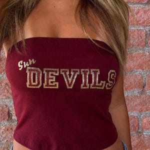 May include: A maroon strapless tube top featuring the text Sun DEVILS printed in a gold glitter finish across the chest. The garment is a fitted, sleeveless style made of a solid-colored fabric, showcasing a clear and bold graphic design.