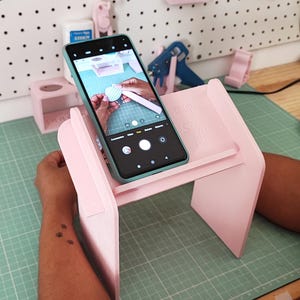 May include: A light pink phone stand holds a teal phone, displaying a crafting project. The stand sits on a green cutting mat. The background includes a pegboard with craft supplies and tools, including scissors and pink accessories.
