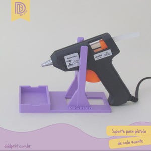 Pode incluir: Uma pistola de cola preta e laranja com uma barra de cola branca está em um suporte roxo. O suporte tem uma pequena bandeja e o texto "dddprint.com.br" é visível. A imagem também inclui o texto "Suporte para pistola de cola quente".