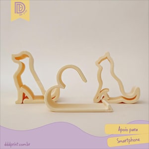 May include: Three beige smartphone holders in the shape of dogs. One is a hanger-style holder, and the other two are dog silhouettes. The text "Apoio para Smartphone" is visible in the image.