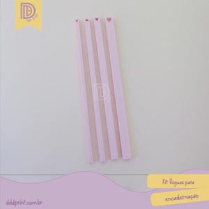 May include: A set of pink and white striped binding rulers with heart-shaped cutouts at the top. The rulers are arranged diagonally against a white background. The image also includes the text "Kit Réguas para encadernação".