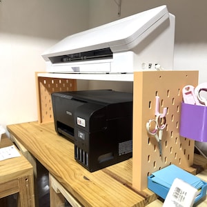 May include: A two-tiered wooden desk organizer with a black printer on the bottom shelf and a white printer on the top shelf. A pegboard with scissors and other craft supplies is on the right.
