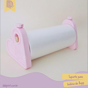 Puede incluir: Un soporte rosa y blanco para rollo Bopp. El soporte tiene un diseño en forma de corazón en los lados y un rollo blanco cilíndrico en el centro. El texto "dddprint.com.br" es visible en la parte inferior.