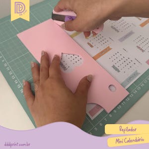 May include: A pink paper calendar sleeve with a heart-shaped cutout is being trimmed with a purple paper cutter. The calendar inside is visible, with months labeled. The text "Refilador" and "Mini Calendário" are visible.