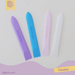 Puede incluir: Cuatro plegaderas de papel de colores: morado, azul, rosa y blanco, cada una con la inscripción "DDDPrint". Están dispuestas sobre una superficie blanca. La imagen también incluye los textos "Dobradeira" y "dddprint.com.br".