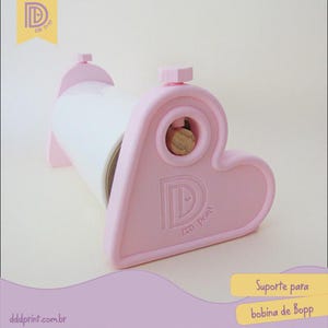 May include: A pink heart-shaped dispenser with a white roll inside. The dispenser has a logo and the text "dddprint.com.br". The text "Suporte para bobina de Bopp" is also visible.