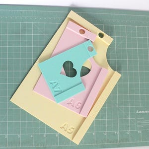 Pode incluir: Um conjunto de ferramentas de artesanato em papel em cores pastel, incluindo uma base amarela com A5, modelos rosa e azul com as marcações A6 e A7 e um recorte em forma de coração. As ferramentas estão dispostas sobre uma base de corte verde.