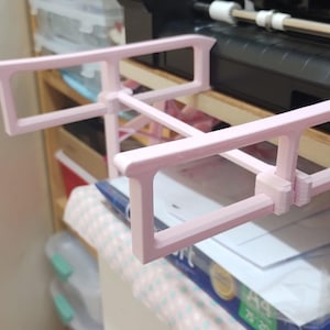 Pode incluir: Uma estrutura retangular rosa claro impressa em 3D com um design semelhante a uma escada. A estrutura está posicionada em frente a uma máquina preta e a uma prateleira de madeira com recipientes de armazenamento. A estrutura é provavelmente um componente ou acessório para a máquina.