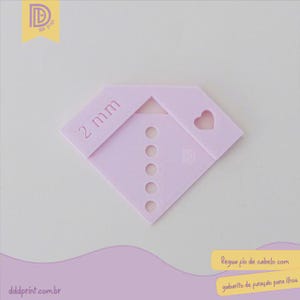 Puede incluir: Una herramienta en forma de diamante rosa claro con el texto "2 mm" y un recorte en forma de corazón. Tiene cinco pequeños agujeros circulares y el logotipo "D". La herramienta se utiliza para hilo de pelo con guía de ojales.