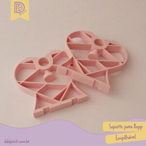 May include: Two light pink, heart-shaped plastic supports with geometric designs. Each has a central circular opening and a screw-like knob. The supports are designed to be stackable, as indicated by the text on the image.