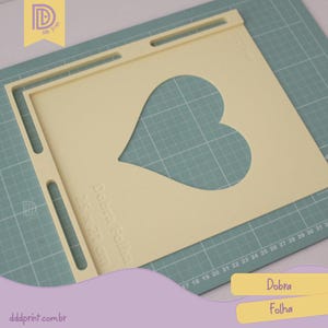 May include: A beige heart-shaped stencil with the text "Dobra Folha 25 x 21 cm" is placed on a green cutting mat. The stencil has a rectangular frame with a heart cutout. The stencil has a handle on the top. The website dddprint.com.br is visible.