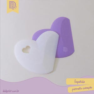 May include: Two heart-shaped spatulas, one white with a heart cutout and one purple. The purple spatula is leaning against the white one. The image has the text "Espátula formato coração" and the website "dddprint.com.br".