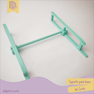 Puede incluir: Un soporte de base de corte verde menta. El soporte tiene una base rectangular con una barra horizontal y un soporte vertical. El texto "dddprint.com.br" es visible en la parte inferior izquierda. El texto "Suporte para base de Corte" es visible a la derecha.