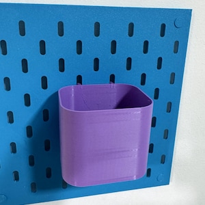 Puede incluir: Un vaso de almacenamiento morado, de forma cuadrada, sujeto a una panel perforado azul. El panel perforado tiene agujeros ovalados espaciados uniformemente. El vaso está hecho de un material sólido y está diseñado para guardar objetos pequeños. El panel perforado está montado en una pared blanca.