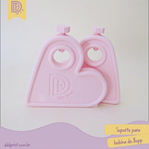 May include: Two light pink, heart-shaped Bopp bobbin holders. Each holder has a circular opening and a small, hexagonal knob at the top. The holders have the text "Ddd Print" and the letter "D" printed on them. The website dddprint.com.br is also visible.