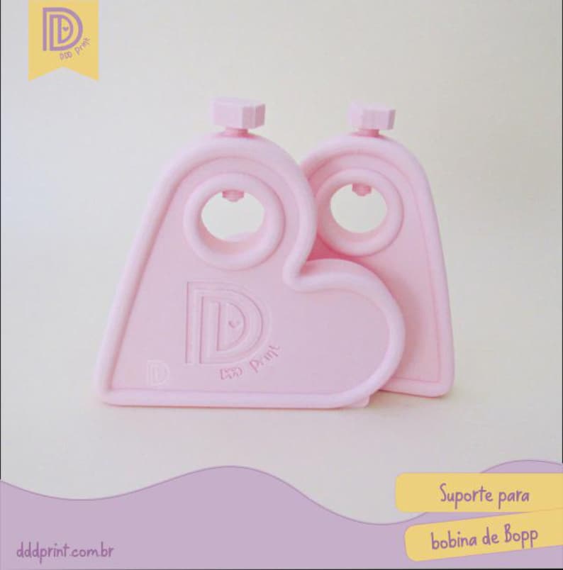 May include: Two light pink, heart-shaped Bopp bobbin holders. Each holder has a circular opening and a small, hexagonal knob at the top. The holders have the text "Ddd Print" and the letter "D" printed on them. The website dddprint.com.br is also visible.